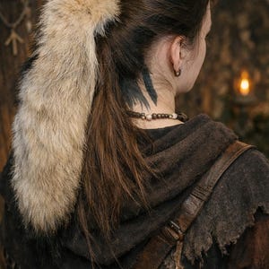May include: A person's back is shown, wearing a brown hooded cloak and a fur hair accessory. The fur is light brown and cream-coloured. A leather strap crosses the shoulder. A tattoo is visible on the neck.