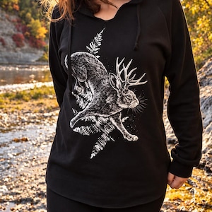 May include: A black long-sleeved hooded sweatshirt featuring a white graphic print of a jackalope leaping through fern fronds. The hoodie has a V-neckline, a drawstring hood, and long sleeves with ribbed cuffs, showcasing a detailed illustration.