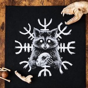 May include: A black fabric square featuring a white graphic print of a raccoon holding a small animal skull. Behind the raccoon is a white circular runic symbol. The design is centered on the fabric, which is displayed on a wooden surface.