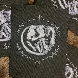 May include: Dark fabric patches with a white illustration of a curled animal inside a circular wreath. The patches have white runes and floral details. The patches are scattered on a brown, textured surface.
