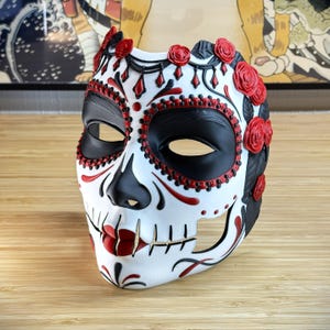 Op de afbeelding: Een wit Day of the Dead-masker met zwarte oogkassen, neus en accenten. Rode rozen en decoratieve elementen sieren het voorhoofd en de randen van het masker. Het masker heeft een half-schedelontwerp met een gedetailleerde, geschilderde afwerking.