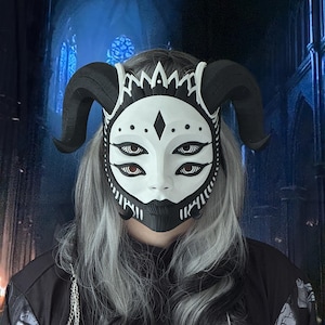 Könnte beinhalten: Eine weiß-schwarze Maske mit vier Augen und großen schwarzen Hörnern. Die Maske hat ein detailliertes Design mit geometrischen Mustern. Ein einzigartiges Accessoire, getragen von einer Person mit grauem Haar.
