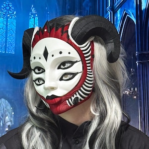 Peut inclure: Un masque blanc à quatre yeux, eye-liner noir et lèvres rouges, doté de grandes cornes noires incurvées sur les côtés. Le masque présente un motif côtelé rouge et blanc autour de la mâchoire et un détail rouge en forme de couronne au sommet.