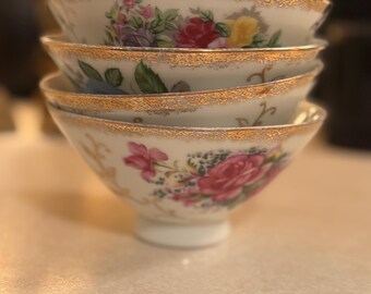 Puede incluir: Una pila de cuatro cuencos de porcelana blanca con diseños florales y ribetes dorados. Los cuencos presentan flores rosas, rojas, amarillas y moradas con hojas verdes. Los bordes están acentuados con oro.