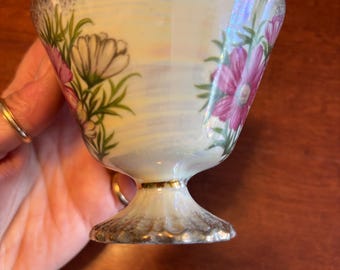 May include: A vintage, iridescent teacup with a pedestal base. The cup is decorated with pink and white floral designs and green leaves. The rim and base are trimmed with gold accents.