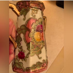 Könnte beinhalten: Vintage, handbemalter Porzellan-Krug mit weißem Grund, verziert mit floralen Designs in Rosa, Gelb und Lila. Der Krug hat goldfarbene Akzente am Griff und am Rand sowie grüne Blätter. Eine kleine rote Rose ist ebenfalls zu sehen.