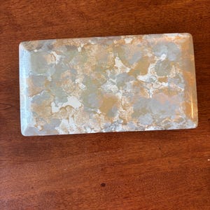 May include: Rectangular decorative box with a glossy finish. The box features a pattern of gold, silver, and white colors. The box is a rectangle with rounded corners. The box is sitting on a wooden surface.
