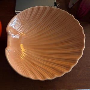 Könnte beinhalten: Eine pfirsichfarbene, keramik Muschelschalenförmige Servierschale. Die Schale hat eine gerippte Textur, die dem Inneren einer Muschel ähnelt. Die Kanten sind gewellt und es gibt dekorative Wirbel an den Seiten. Diese dekorative Schale eignet sich perfekt zum Servieren von Vorspeisen oder zum Präsentieren von Dekorationsartikeln.