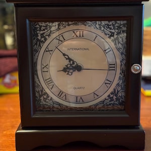 Puede incluir: Un reloj de cuarzo de madera marrón oscuro con una esfera cuadrada. La esfera del reloj es de color crema con números romanos negros y la palabra "INTERNATIONAL" encima de las manecillas. El reloj tiene un asa decorativa plateada en la parte superior y un botón plateado en el lateral.