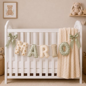 Op de afbeelding: Een wit ledikant met een decoratieve slinger van gevulde stoffen letters die het woord MARIO vormen. De letters hebben diverse patronen zoals teddyberen, groen ruitjesmotief en sterren. Twee saliegroene satijnen strikken sieren de uiteinden.