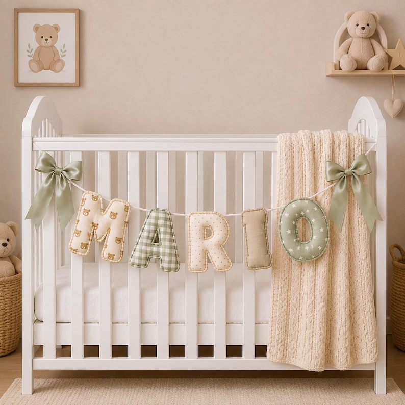 Op de afbeelding: Een wit ledikant met een decoratieve slinger van gevulde stoffen letters die het woord MARIO vormen. De letters hebben diverse patronen zoals teddyberen, groen ruitjesmotief en sterren. Twee saliegroene satijnen strikken sieren de uiteinden.