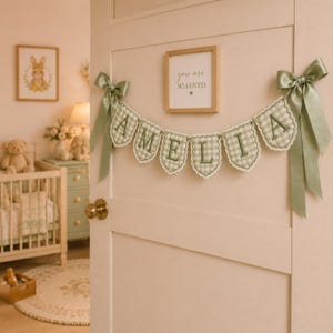 May include: A decorative fabric banner featuring seven scalloped pennants in a light green and white gingham pattern. Each pennant displays a dark green embroidered letter spelling AMELIA. The banner is accented with sage green satin bows.