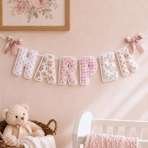 Op de afbeelding: Een stoffen slinger die de naam HARPER spelt in individuele gevulde letters. Elke letter heeft een ander roze en wit patroon, waaronder hartjes, bloemenprints en vichy-ruitjes, afgewerkt met een witte schulprand en roze strikjes.