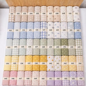 Op de afbeelding: Een verzameling stofstalen gerangschikt in een raster, elk gelabeld met de nummers 101 tot en met 155. De textielsoorten bevatten diverse patronen zoals strepen, vichy-ruiten, stippen, wolken, sterren, bloemen en kleine diermotieven.