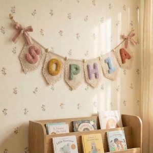 Op de afbeelding: Een stoffen slinger met de naam SOPHIA op individuele wimpels. Elke wimpel is gemaakt van crèmekleurige stof met kanten randen en een getextureerde, getufte letter in roze, geel, groen, paars, blauw en oranje.