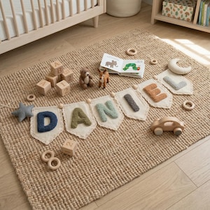 Op de afbeelding: Een kinderkamerscène met een jute vloerkleed en een banner met de naam "DANI" in getextureerde letters. Houten blokken, een teddybeer, een hertfiguur en een houten auto zijn erop geplaatst. Een kinderboek en een stervormige decoratie zijn ook zichtbaar.