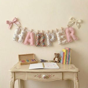 Op de afbeelding: Een stoffen slinger die de naam Harper spelt met gevulde, gedessineerde letters die aan een lint hangen met houten clips en crèmekleurige strikken aan beide uiteinden. De letters hebben bloemen- en ruitjesprints in roze, blauw en wit.