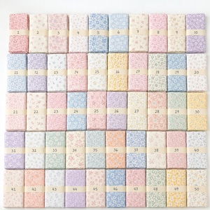 Op de afbeelding: Een verzameling van vijftig rechthoekige stoffen stalen gerangschikt in een raster, elk met een uniek bloemenpatroon in pasteltinten van roze, blauw, geel, groen en paars. Elke staal is vastgezet met een beige papieren band gelabeld van 1 tot 50.