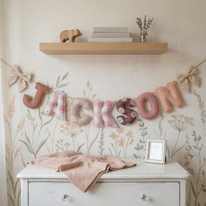 May include: A decorative fabric banner spelling the name JACKSON in individual stuffed letters. The letters feature various patterns including pink gingham, solid dusty rose, and brown with white daisy prints, finished with beige polka dot bows.