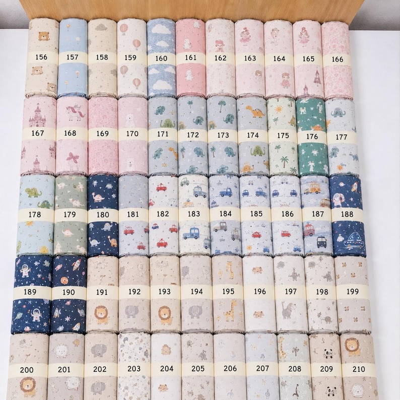 Op de afbeelding: Een collectie stoffen rollen gerangschikt in een raster, elk met unieke patronen waaronder dieren, voertuigen, ruimtethema's en kastelen. Elke rol is genummerd van 156 tot 210 en toont een verscheidenheid aan pastelkleuren en opvallende kleuren.