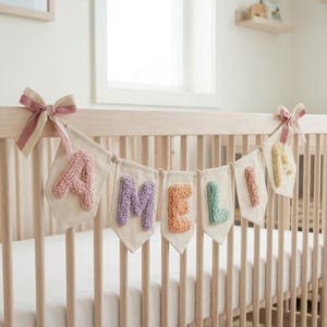 Op de afbeelding: Een banner met de naam "AMELIA" hangt boven een wieg. De letters zijn in verschillende pastelkleuren en hebben een getextureerde, getufte uitstraling. De banner is gemaakt van ecru stof en is vastgebonden met roze strikken.