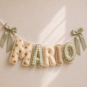Op de afbeelding: Een decoratieve stoffen slinger die de naam MARIO spelt in gevulde, zachte letters. De letters hebben diverse patronen zoals bruine teddyberen, groen ruitjespatroon, beige stippen, effen beige en groen met witte sterren, afgewerkt met saliegroene strikken.