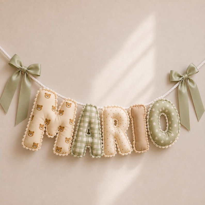 Op de afbeelding: Een decoratieve stoffen slinger die de naam MARIO spelt in gevulde, zachte letters. De letters hebben diverse patronen zoals bruine teddyberen, groen ruitjespatroon, beige stippen, effen beige en groen met witte sterren, afgewerkt met saliegroene strikken.