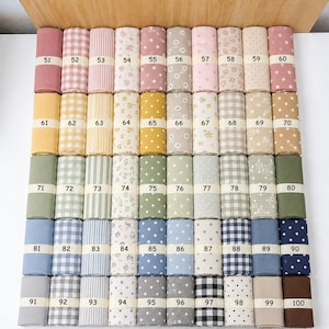 Op de afbeelding: Een collectie van vijftig stofrollen gerangschikt in een raster, elk omwikkeld met een papieren band met de nummers 51 tot en met 100. De stoffen bevatten diverse patronen, waaronder strepen, ruitjes, stippen, sterren en bloemenprints.