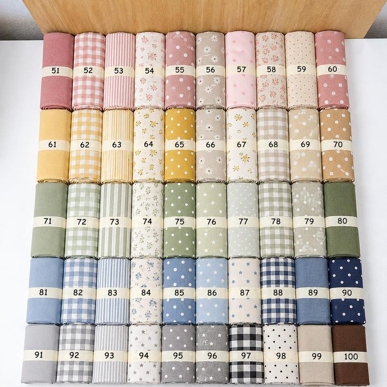Op de afbeelding: Een collectie van vijftig stofrollen gerangschikt in een raster, elk omwikkeld met een papieren band met de nummers 51 tot en met 100. De stoffen bevatten diverse patronen, waaronder strepen, ruitjes, stippen, sterren en bloemenprints.