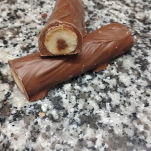 May include: Two chocolate-covered candy bars, one cut in half to reveal a light-colored filling with a dark center. The bars are on a speckled gray surface. The chocolate coating is a rich brown color.