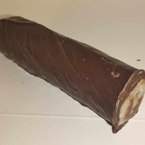 Puede incluir: Un dulce largo y cilíndrico cubierto de chocolate. El exterior marrón oscuro tiene un aspecto texturizado, y el extremo cortado revela un relleno de color claro. El dulce está colocado en diagonal sobre un fondo blanco.