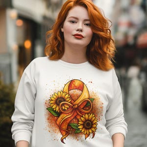 Pode incluir: Camisola branca com um desenho em aquarela de uma fita amarela, girassóis e um laço. O desenho está centrado na frente. A modelo está vestindo calças cinza e está em uma rua de paralelepípedos.