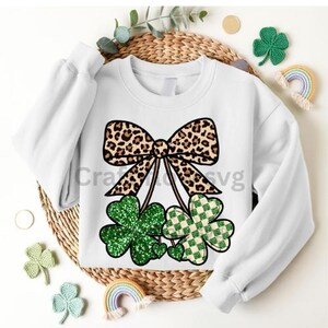 May include: White sweatshirt featuring a leopard print bow and three shamrocks. Two shamrocks are green with glitter, and one is a green and white checkered pattern. The text "Crafts svg" is visible on the sweatshirt.