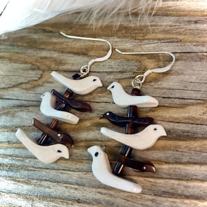 May include: A pair of dangle earrings featuring three white bird charms stacked on top of each other, with brown accents. The earrings are made with silver hooks.
