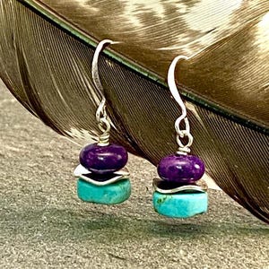 May include: A pair of handcrafted earrings with a layered design. Each earring features a turquoise stone at the base, a silver accent, and a deep purple bead at the top. The earrings hang from silver hooks.