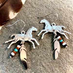 May include: Silver-toned horse earrings with dangling feathers and beaded accents. The horse pendants have a running pose and are detailed with small dots. The feathers are white and black. The earrings have a hook closure.