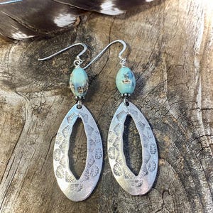 May include: Silver-toned dangle earrings with a teardrop shape and floral pattern. Each earring features a light blue, oval-shaped bead. The earrings are suspended from silver-colored ear wires. The background is a wooden surface.