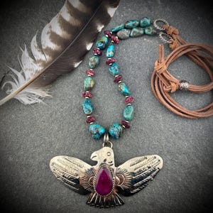 May include: A turquoise and red beaded necklace with a silver eagle pendant. The pendant features a large, teardrop-shaped red stone. A brown leather cord and a feather are also visible.