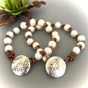 May include: Two pearl bracelets with brown leather cords and silver-tone clasps. Each bracelet features a series of white pearls strung together with knotted brown leather. The clasps are round with a floral design.