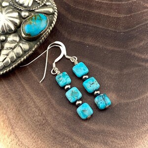 May include: Dangle earrings featuring three square-cut turquoise stones separated by small silver-toned metal beads on each earring. The earrings have silver-toned ear wires and are positioned next to a silver-toned metal object with a turquoise inlay.