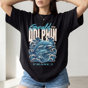 Może przedstawiać: Czarny t-shirt z grafiką przedstawiającą dwa delfiny wyskakujące z niebieskich fal. Tekst "Gerald the Dolphin" znajduje się nad obrazem, a pod nim "PHASE 2". Projekt wykorzystuje odcienie niebieskiego i białego.