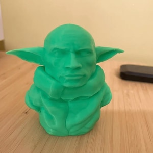 Pode incluir: Uma estatueta verde impressa em 3D de um personagem com orelhas grandes e uma estrutura facial distinta. A figura é um busto, com um rosto detalhado e uma roupa texturizada e em camadas. A estatueta é exibida em uma superfície de madeira.