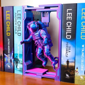 Op de afbeelding: Een 3D-geprinte astronauten boekensteun in blauwe en roze tinten. De astronaut staat in een ruimtestation-achtig frame, met boeken van Lee Child zichtbaar. De boekensteun voegt een unieke, ruimtelijke touch toe.
