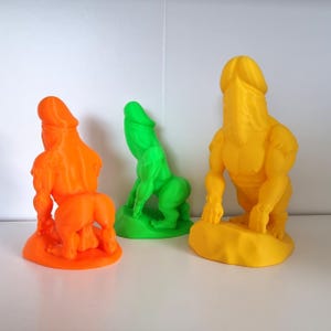 May include: Three brightly coloured sculptures of a male figure with a phallic head and muscular body. The sculptures are in orange, green, and yellow. The figures are posed in a standing position.