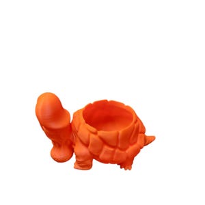 May include: An orange turtle-shaped planter. The planter has a large opening for plants and a detailed shell design. The turtle's head and legs are also visible, creating a whimsical and decorative piece for home decor.