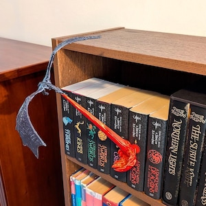 Pode incluir: Um marcador de livros em forma de dragão cinza escuro com detalhes de chama vermelha e laranja, colocado em uma estante. As asas do dragão se estendem sobre os livros, criando um tema de fantasia.