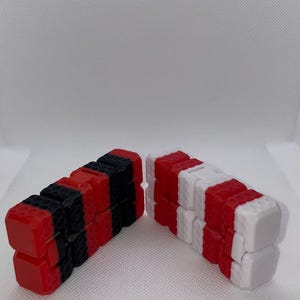 Pode incluir: Dois brinquedos fidget são exibidos. Um é vermelho e preto, o outro é vermelho e branco. Cada brinquedo é feito de blocos retangulares conectados. Os blocos têm uma superfície texturizada. O fundo é branco.