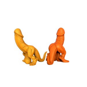 Könnte beinhalten: Zwei neuartige Figuren in Form einer stilisierten, echsenartigen Kreatur. Eine ist goldgelb, die andere orange. Beide haben eine phallische Form und sind auf weißem Hintergrund platziert.