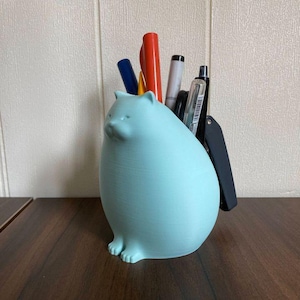 Op de afbeelding: Een lichtblauwe katvormige bureau-organizer met pennen en een briefopener. De organizer is gemaakt van een glad materiaal en heeft een afgerond lichaam met kleine pootjes. De pennen zijn rood, blauw, zwart en zilver.