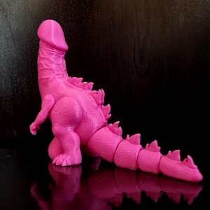 Peut inclure: Figurine rose imprimée en 3D ressemblant à Godzilla, avec un design phallique. La figurine a une surface texturée, une longue queue et des nageoires dorsales. Le jouet est posé sur une surface noire réfléchissante.
